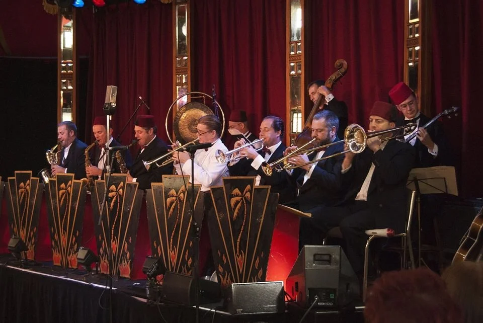 Big band jazz musicians playing instruments on stage, with red curtains in the background.