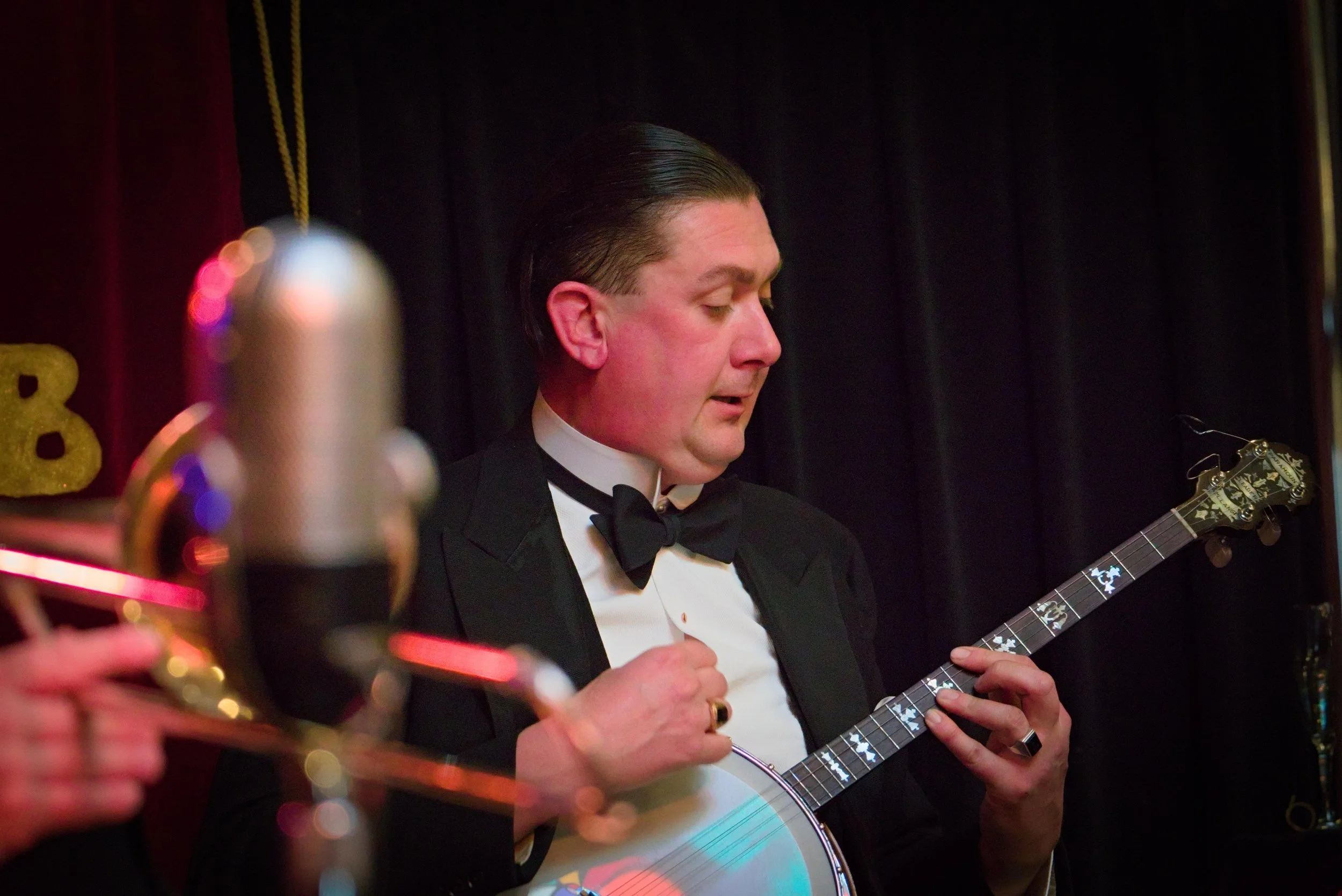 A man in a tuxedo with a bow tie playing an acoustic guitar with a black background.