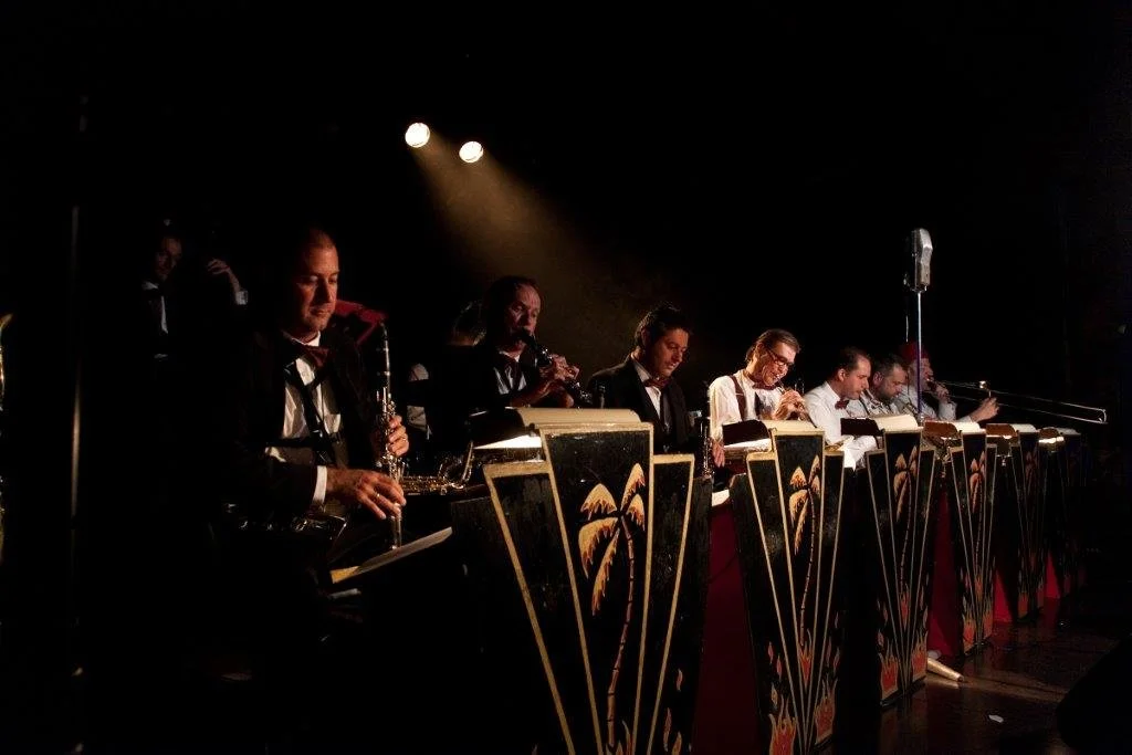 Big band orchestra performing on stage with music stands decorated with palm trees, illuminated by stage lights.