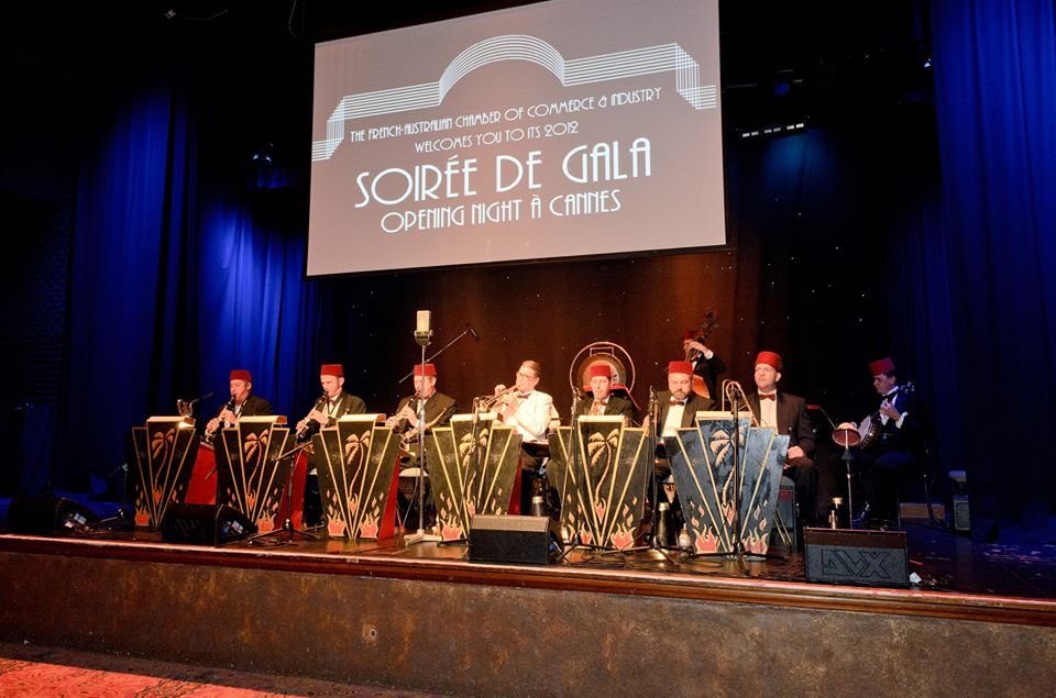 Musicians performing on stage at an event with a sign that reads 'Soirée de Gala Opening Night at Cannes,' organized by the French-Australian Chamber of Commerce & Industry in 2012, with dark blue curtains and stage lighting.