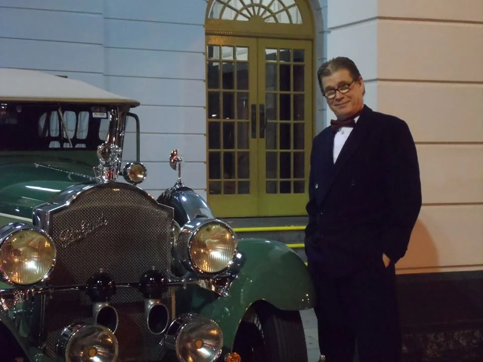 Man in tuxedo standing next to a vintage green car outside a building with yellow double doors.