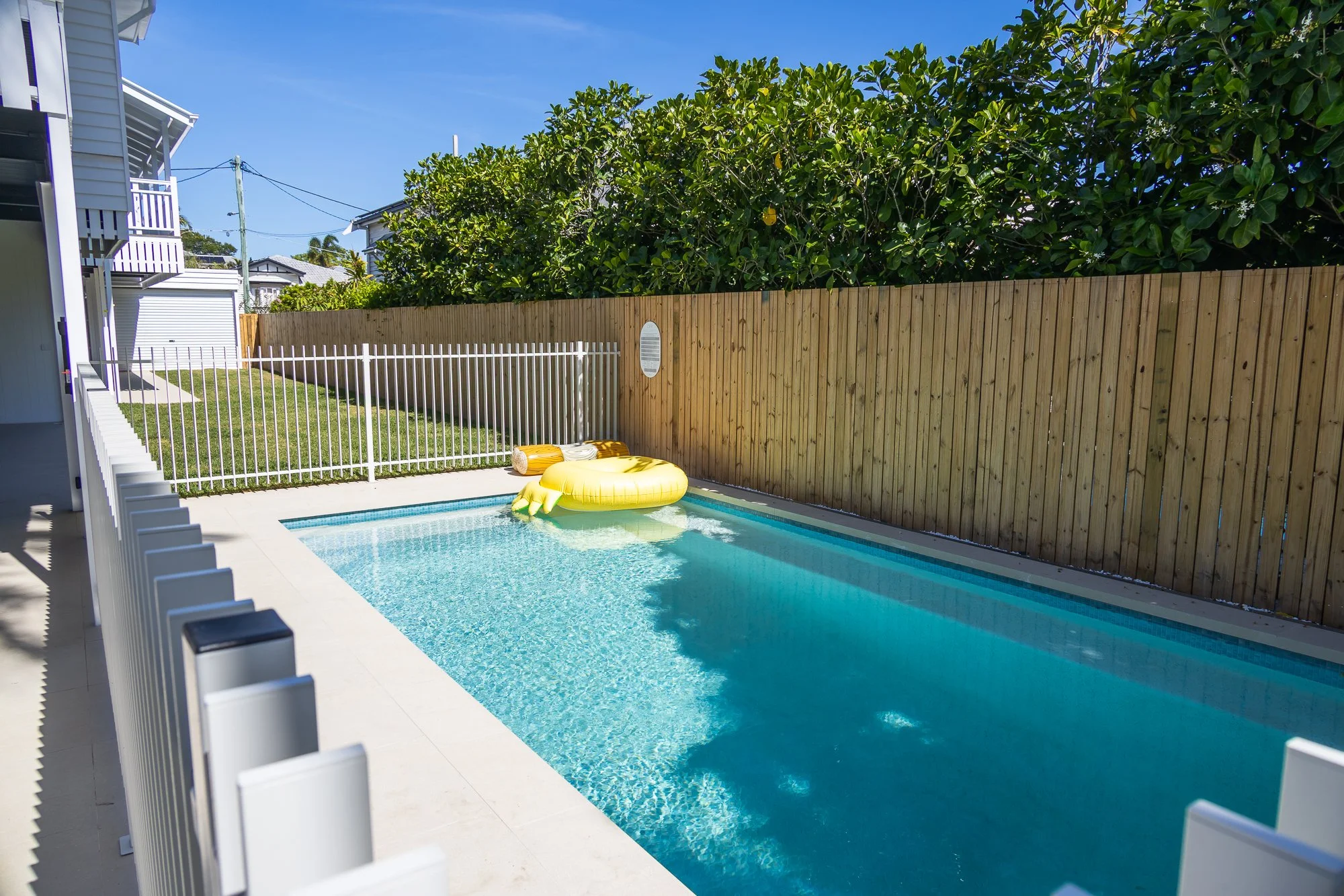 Backyard swimming pool with a yellow inflatable float, surrounded by a white fence and a wooden privacy fence, with green trees and a playground toy in the yard.