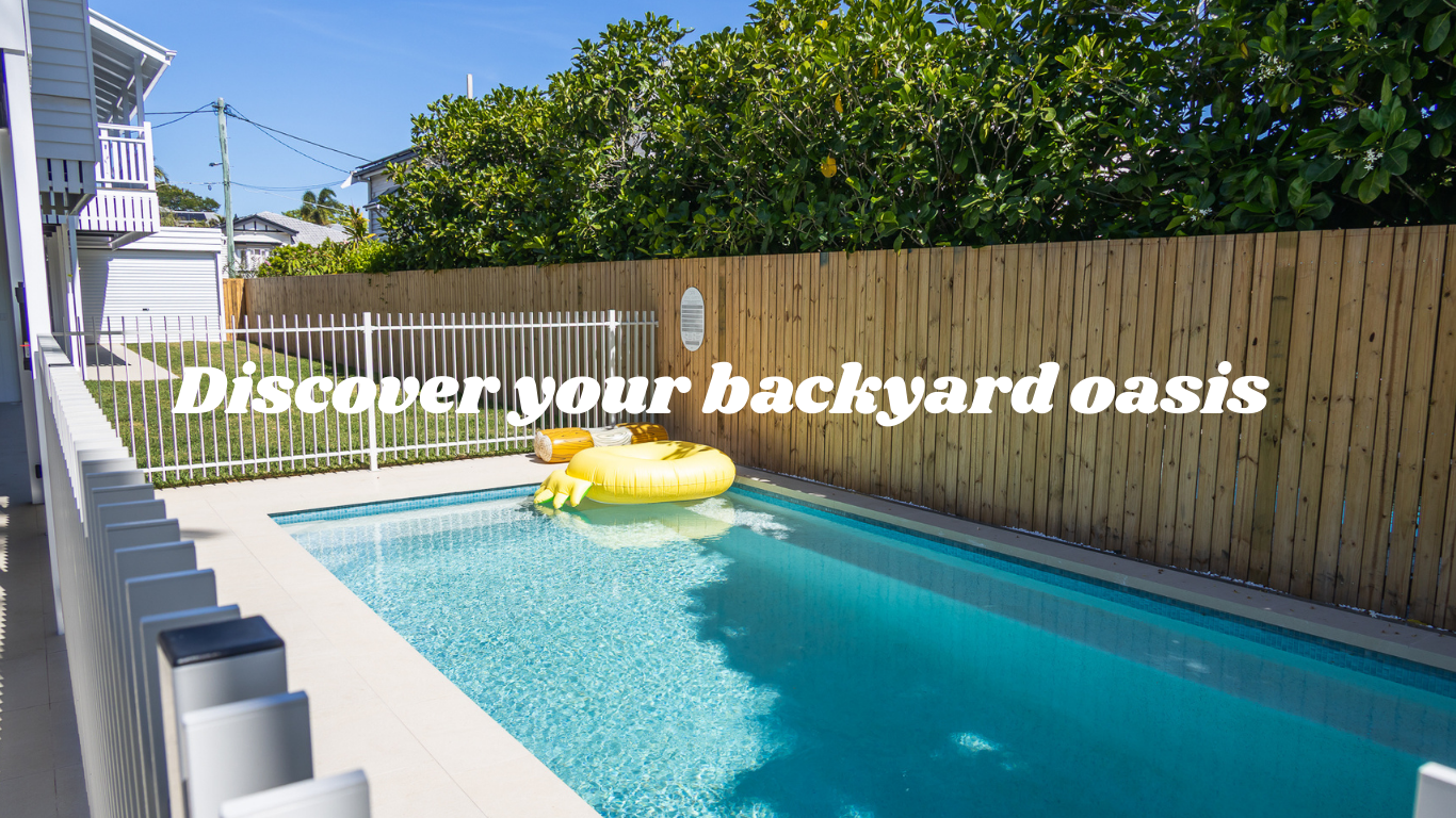 A backyard swimming pool with a yellow inflatable rubber duck float, surrounded by a white fence and a wooden privacy wall, with lush green trees and a clear blue sky above.