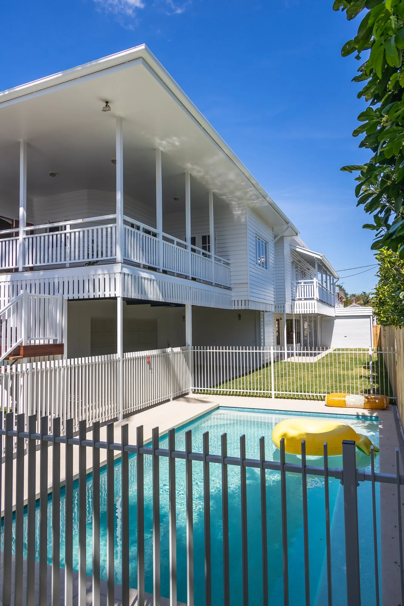Backyard view of a white two-story house with a swimming pool, fenced in with a white metal fence, and a yellow inflatable float in the pool. Bright blue sky above.