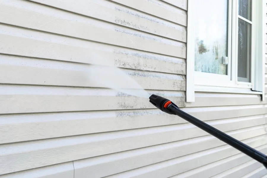 A person pressure washing the siding of a house near a window.