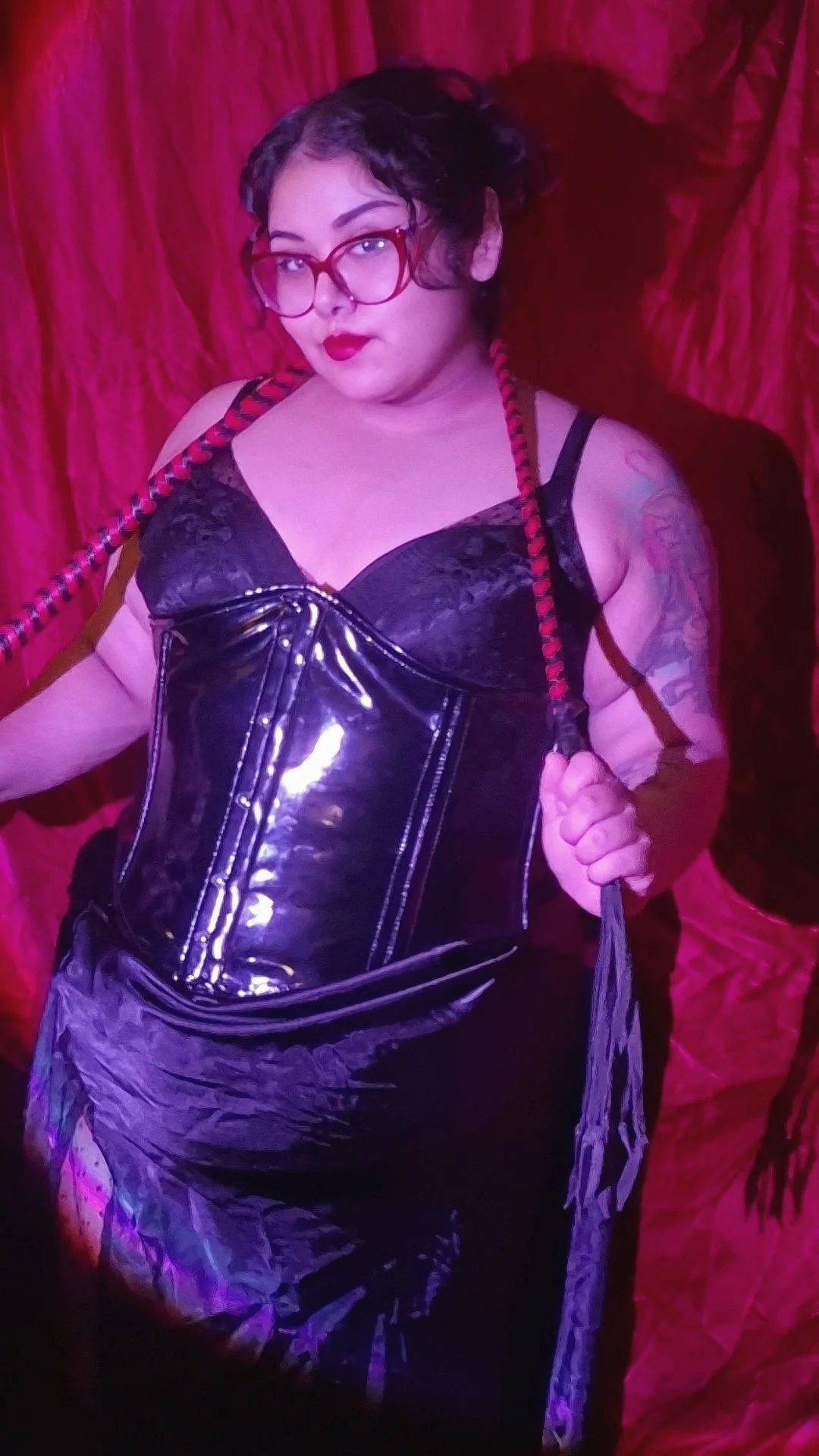A woman with glasses and dark hair styled in braids, wearing a black corset, lace top, and shiny pants, holding a whip, standing against a red curtain background.