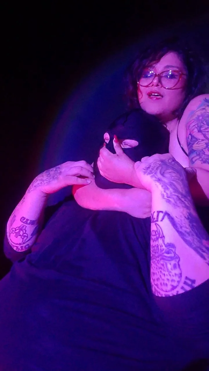 A woman with curly hair and glasses embraces a person wearing a black hoodie, in a setting with purple and blue lighting.