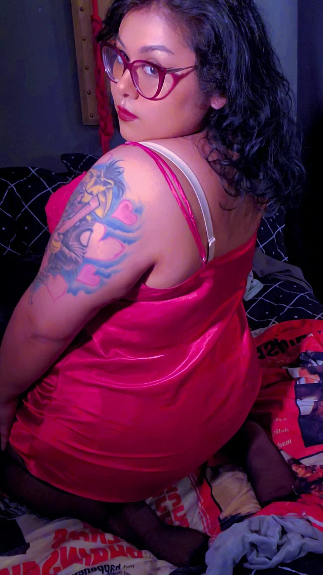 A woman with black curly hair, red glasses, and red lipstick, shows her shoulder tattoo of a dragon and hearts, while wearing a pink satin garment, sitting on a bed with black and white bedspread.