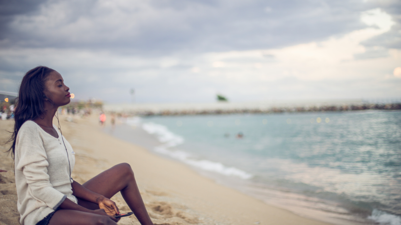 A woman sits on a California beach with her eyes closed, taking a slow breath — pausing in a moment of stillness before returning to the demands of daily life.