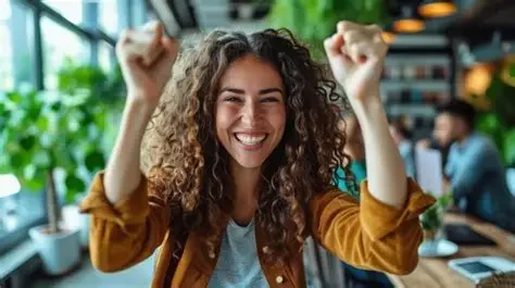 A woman with curly hair smiling and raising her fists in celebration inside a modern indoor space with greenery and people in the background.