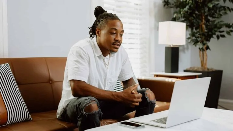 A young man sitting on a brown sofa, looking at a laptop on a white table, with a smartphone nearby. The room has a modern decor with a lamp and a green plant in the background.