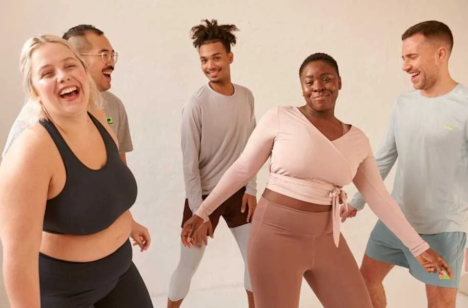 Group of five people standing together, smiling and laughing against a plain wall.
