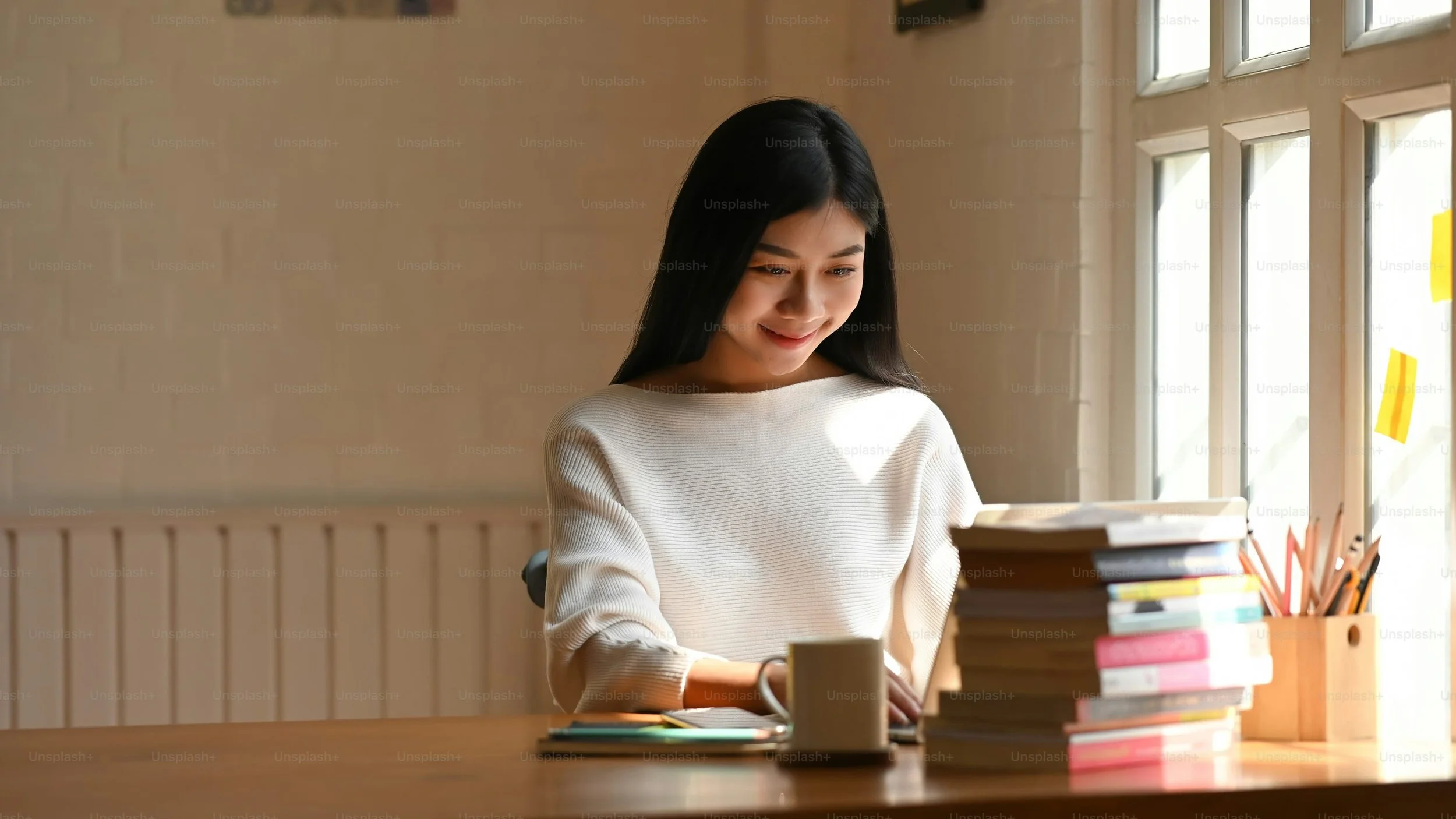 A young woman with long black hair wearing a white sweater sits at a wooden desk with a stack of books, a coffee mug, and writing supplies, smiling as she uses a laptop near a window.