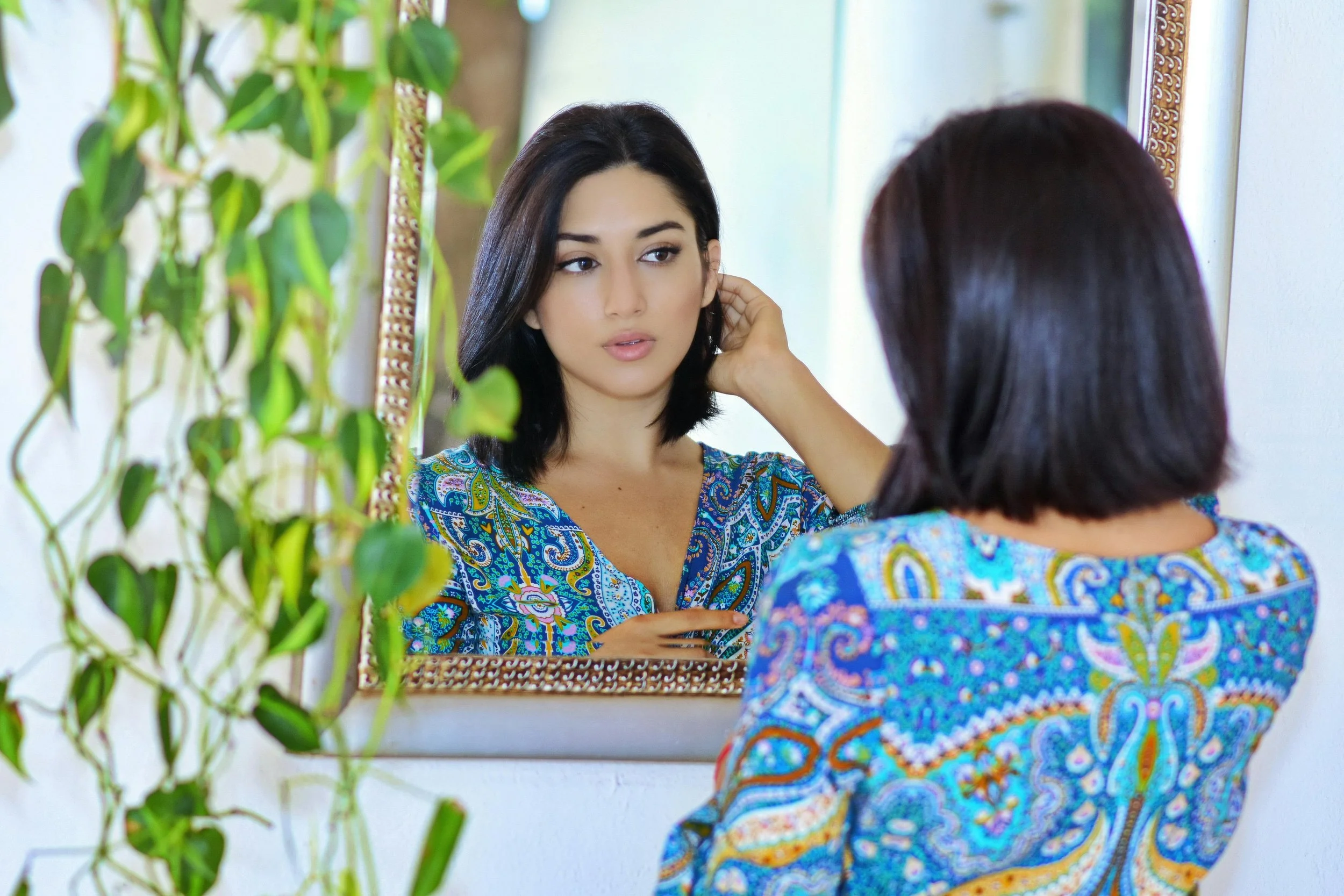 A woman stands at a mirror adjusting her hair, her expression uncertain — a quiet illustration of the intrusive doubt and compulsive checking that characterizes Body Dysmorphic Disorder.