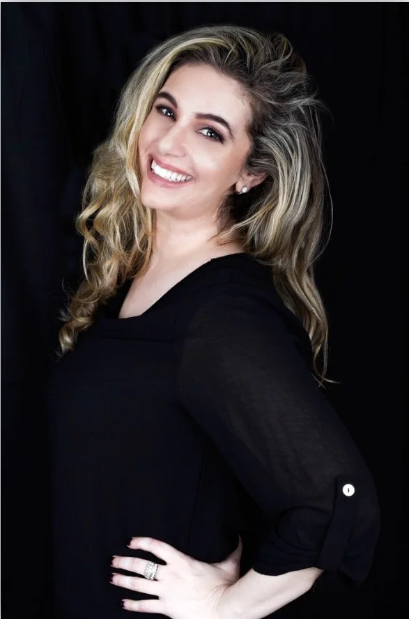 A woman with long, wavy blonde hair, smiling, wearing a black top, standing against a black background.