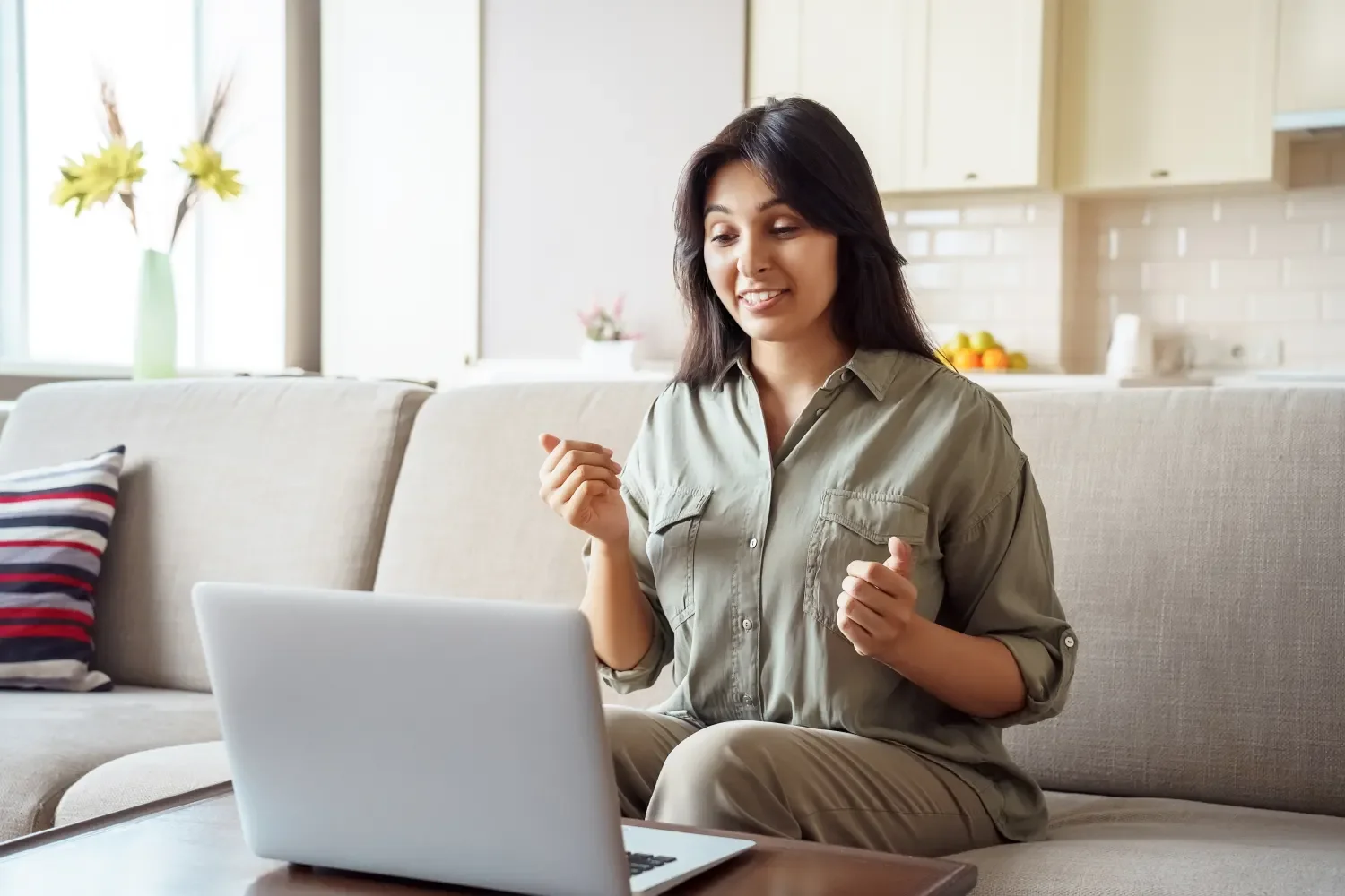 A woman sitting on a beige couch with a laptop in front of her, smiling and gesturing excitedly in a bright living room with a kitchen in the background.