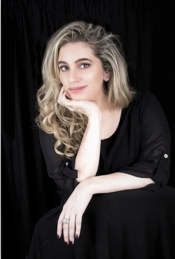 A woman with blonde, curly hair sitting in a black outfit against a dark background, resting her chin on her hand and looking at the camera.
