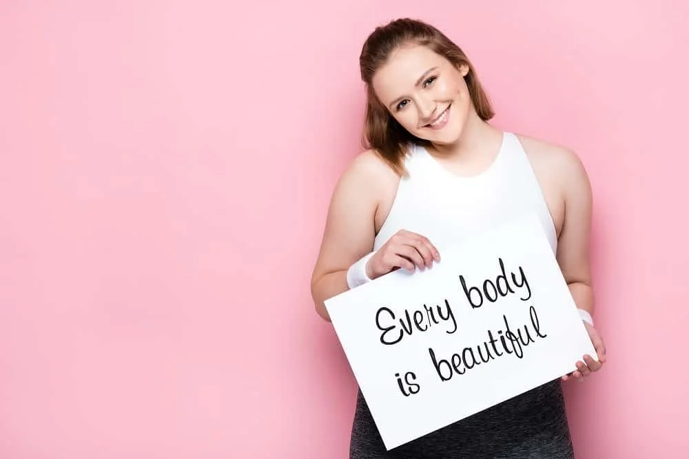 A young woman with a big smile holding a sign that says 'Every body is beautiful' against a pink background.