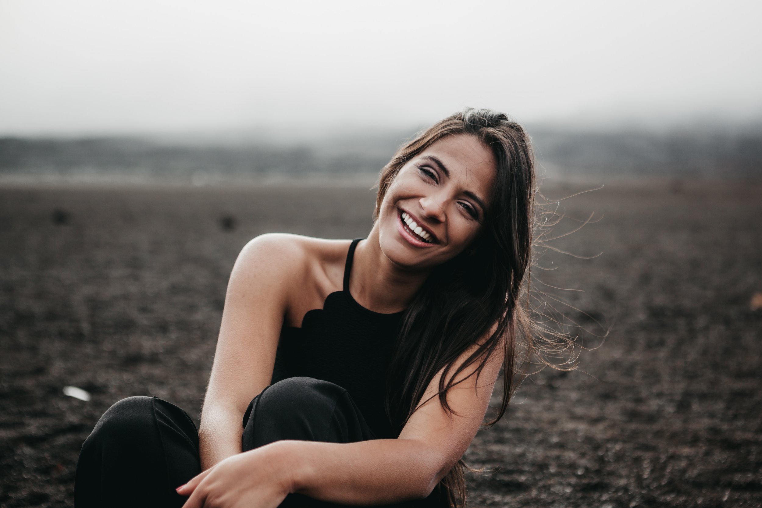 Smiling woman sitting on dark soil or volcanic ash, outdoors, with a blurred landscape background and overcast sky.