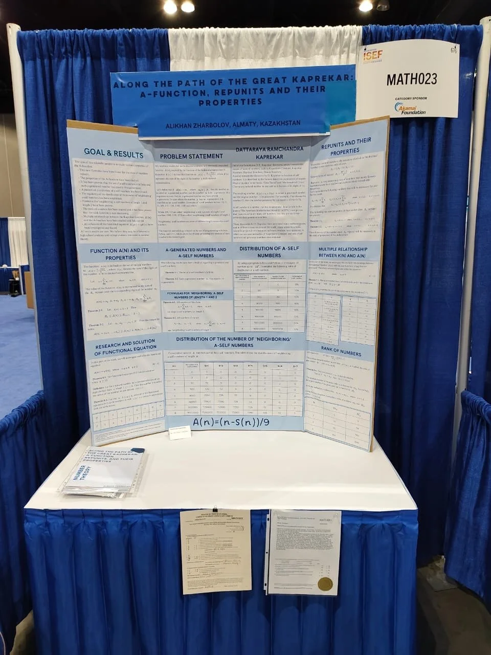 Academic research poster titled 'Along the Path of the Great Kaprekar: A-Function, Repunits and their Properties' by Alikhan Zharbolov from Almaty, Kazakhstan, displayed on a booth at a conference with blue curtains. The poster contains sections with formulas and tables related to mathematical properties of Kaprekar numbers.