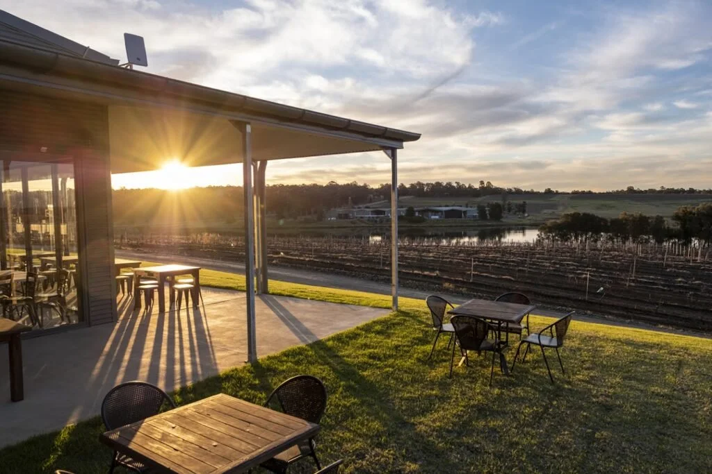 Outdoor patio area with tables and chairs, overlooking a vineyard and a small body of water at sunset, partially covered by a roof.