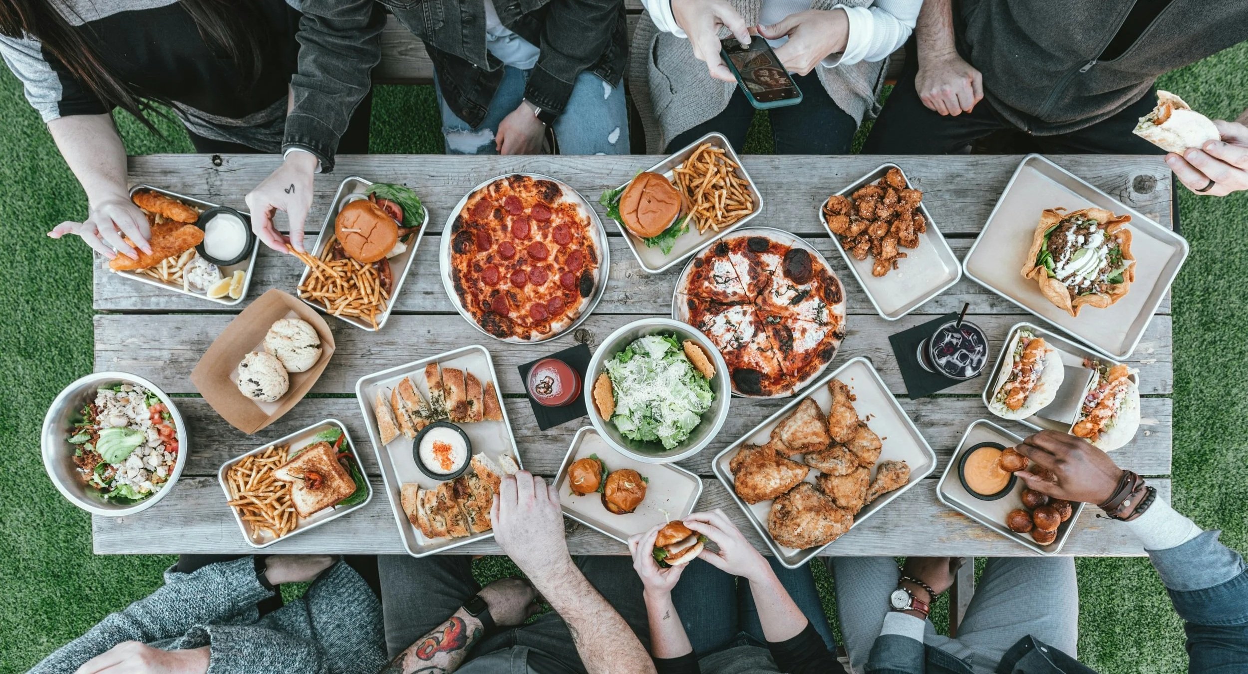 A top-down view of a group of people having a meal outdoors at a wooden picnic table. The table is filled with various dishes including pizza, salads, sandwiches, fried chicken, French fries, meats, and drinks.