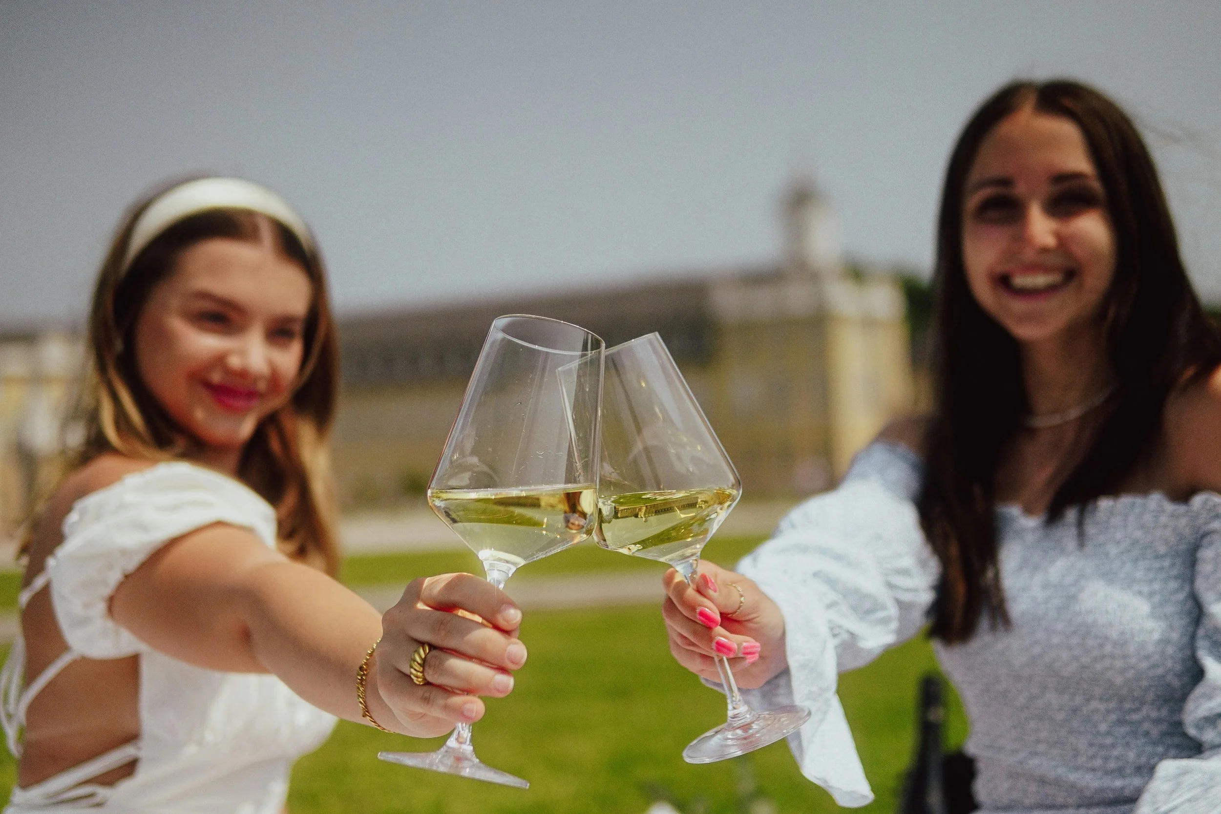 Two women toasting with glasses of white wine outdoors, smiling, with a historic building in the background.