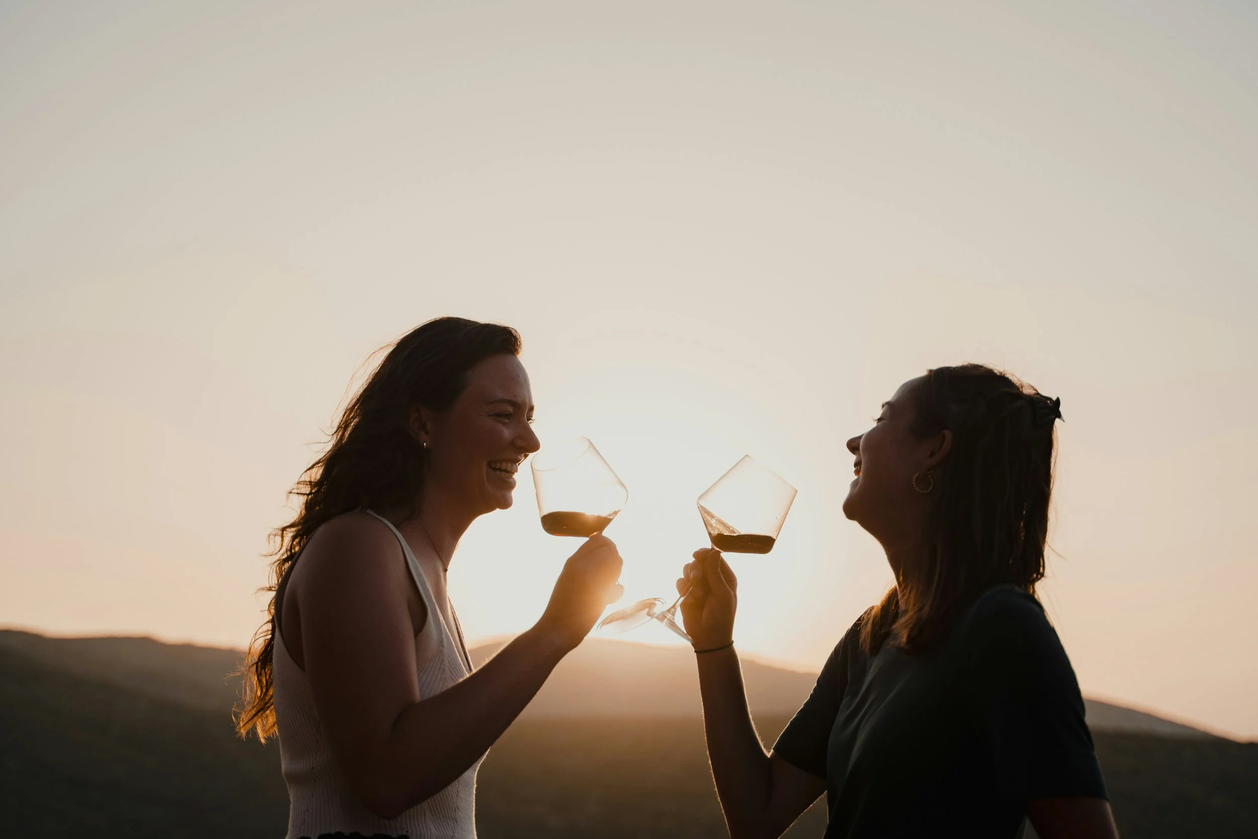 Two women smiling and holding glasses of wine during sunset, with a hilly landscape in the background.