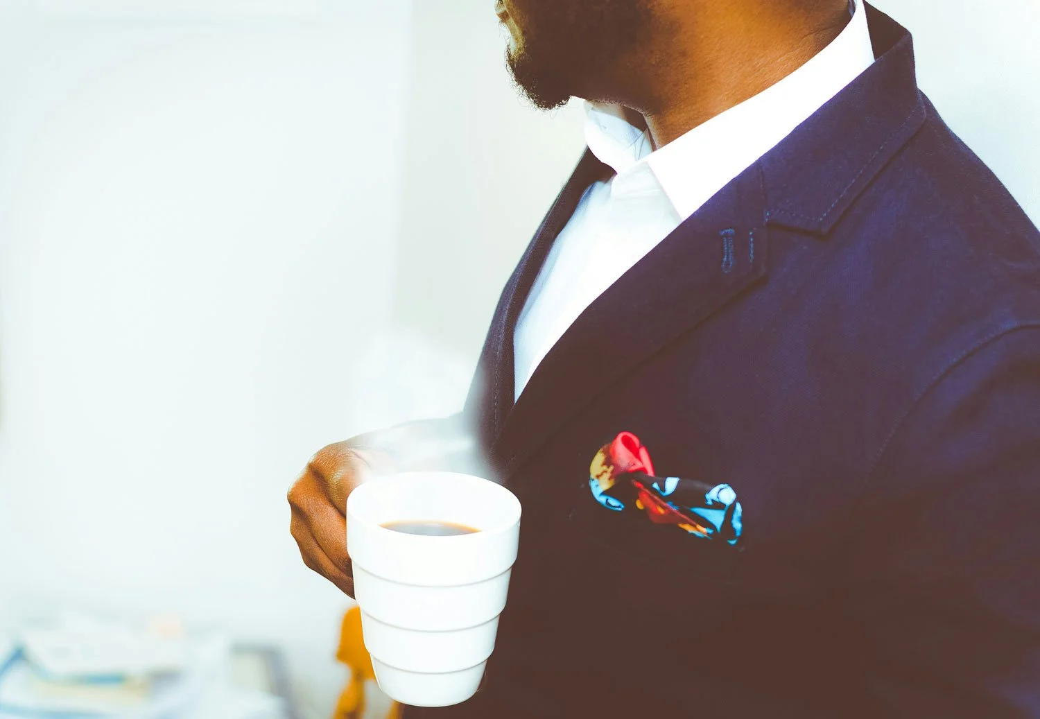 Close-up of a man wearing a dark suit with a colorful pocket square, holding a steaming cup of coffee.