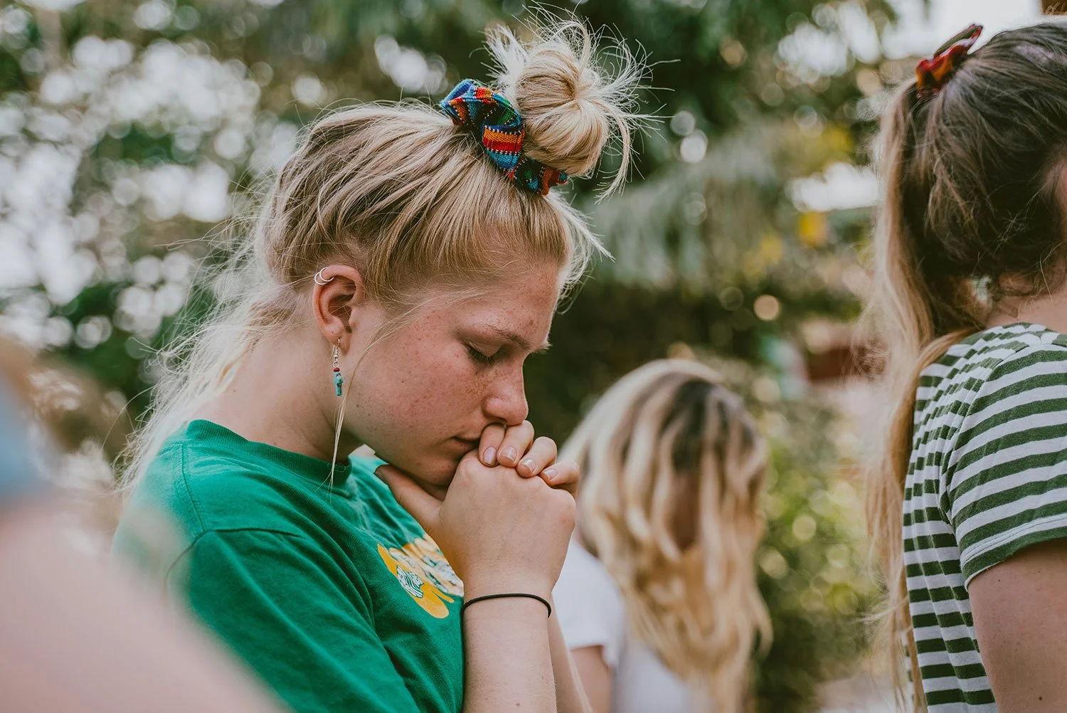 A young girl with blonde hair tied up with a colorful scrunchie, wearing a green t-shirt, closed eyes, and prayerful posture outdoors surrounded by other people.