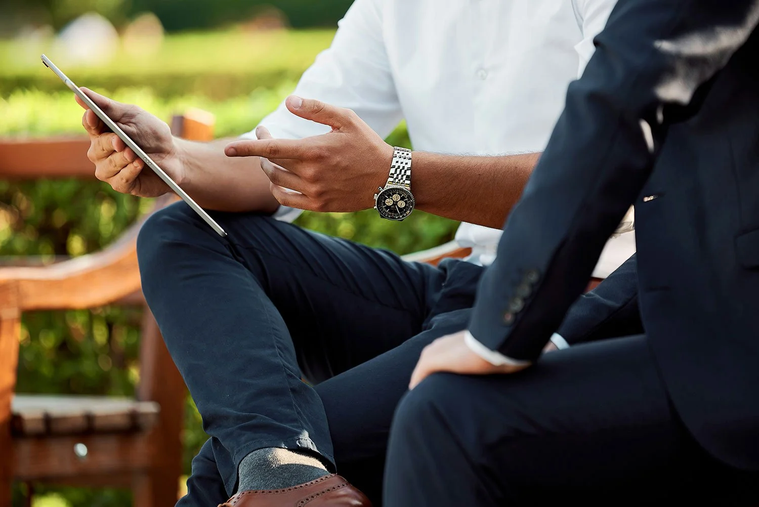 Two men in formal attire sitting outdoors, one showing something on a tablet to the other.