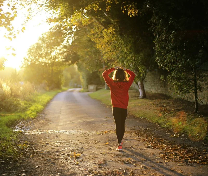 A woman walking down a sunlit country road during autumn, wearing a red jacket and black pants, with trees lining the road.