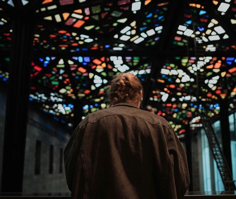 A person with braided hair wearing a brown jacket, seen from behind, looking up at a colorful, stained-glass ceiling.