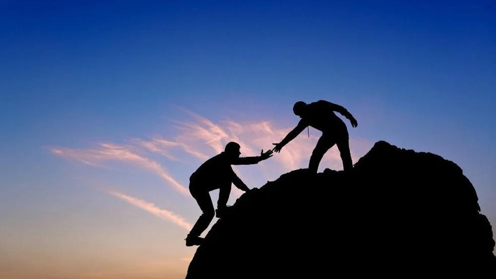 Silhouettes of two people on a mountain, one helping the other to climb up during sunset.