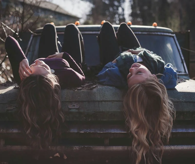 Two young women lying on the hood of a truck, looking up and relaxing outdoors with hilly landscape in the background.