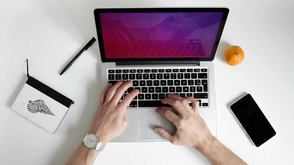A person working on a silver laptop at a white desk, with a black pen, a small notebook with a shell drawing, an orange, and a black smartphone on the desk.