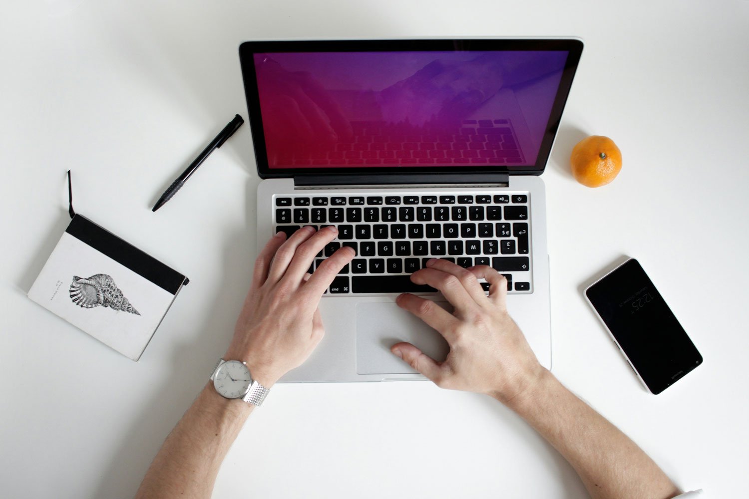 Overhead view of a person typing on a laptop with various items on a white desk, including a black pen, a notepad with a seashell drawing, a tangerine, and a smartphone.