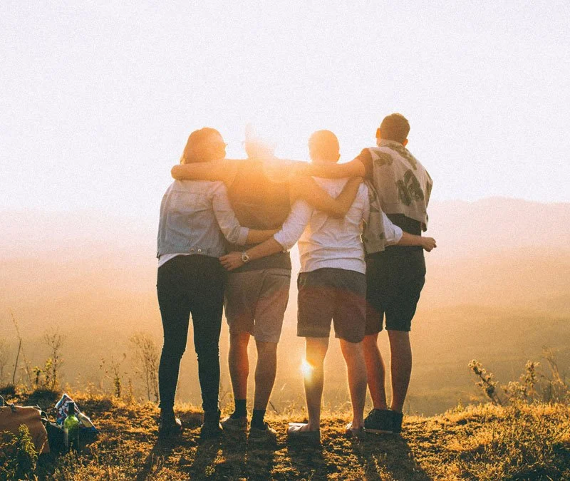 Four friends standing with arms around each other at sunset, overlooking a scenic landscape.
