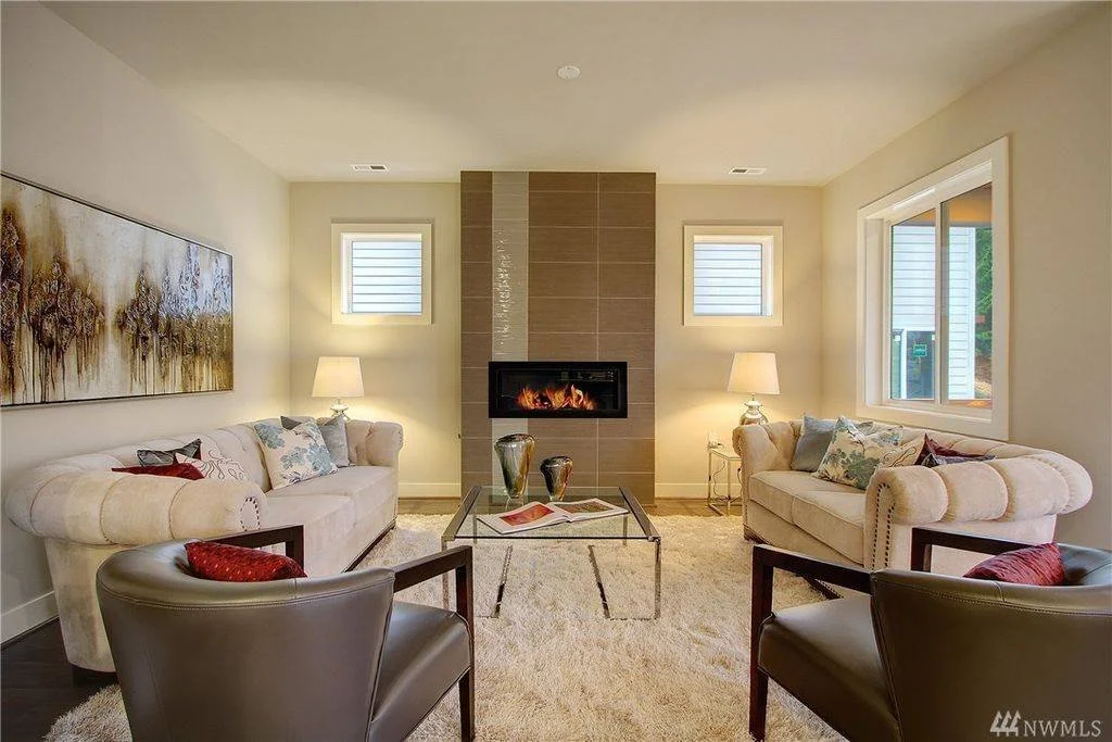 Living room with two beige sofas, two dark brown armchairs, a glass coffee table, and a modern fireplace with a tile surround. There are two small windows above the sofas, two table lamps on side tables, artwork on the wall, and a large window to the