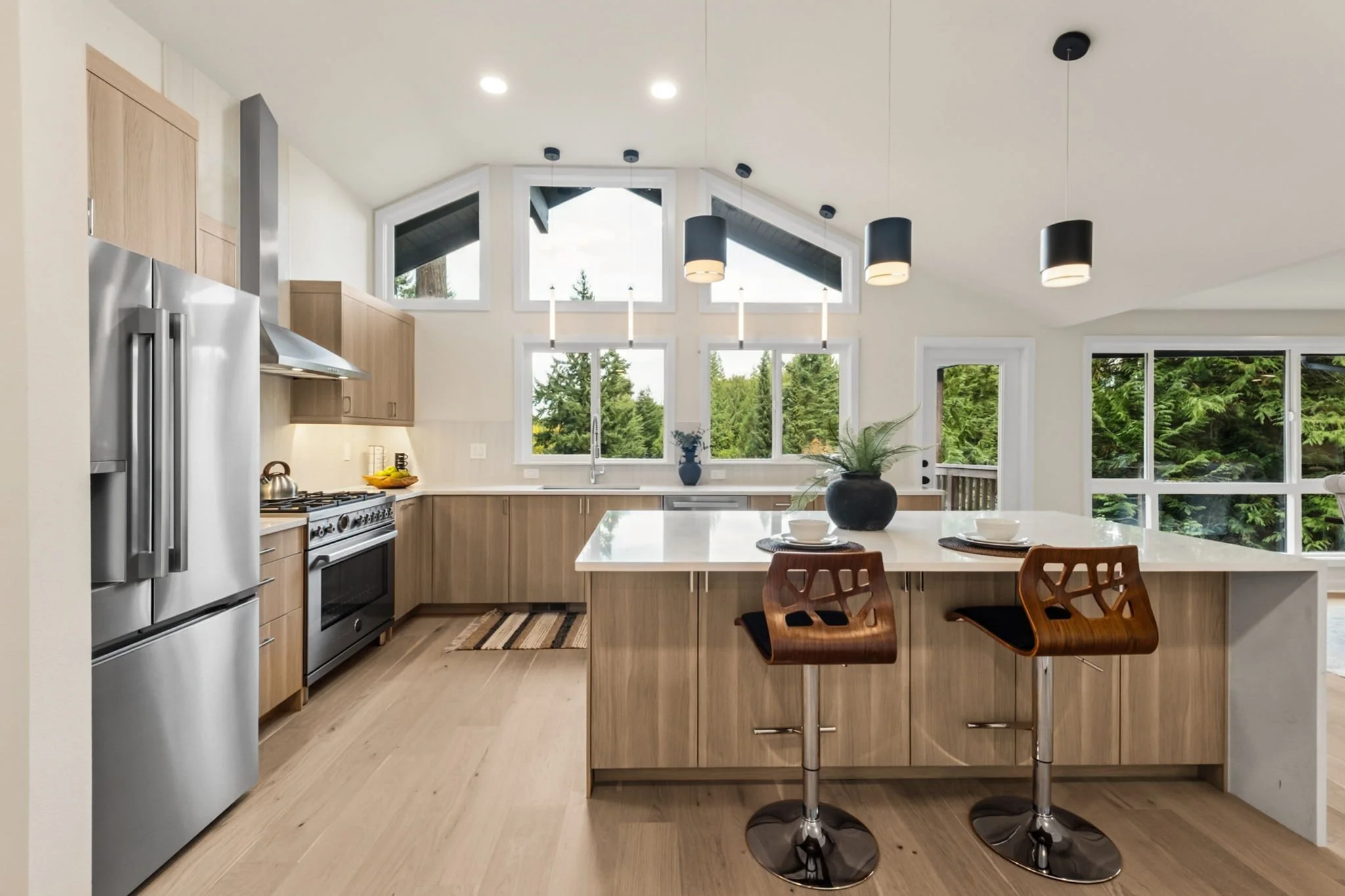 Modern kitchen with wooden cabinets, stainless steel refrigerator and stove, white countertops, large window overlooking trees, and two wooden bar stools at a white island with decorative vases.