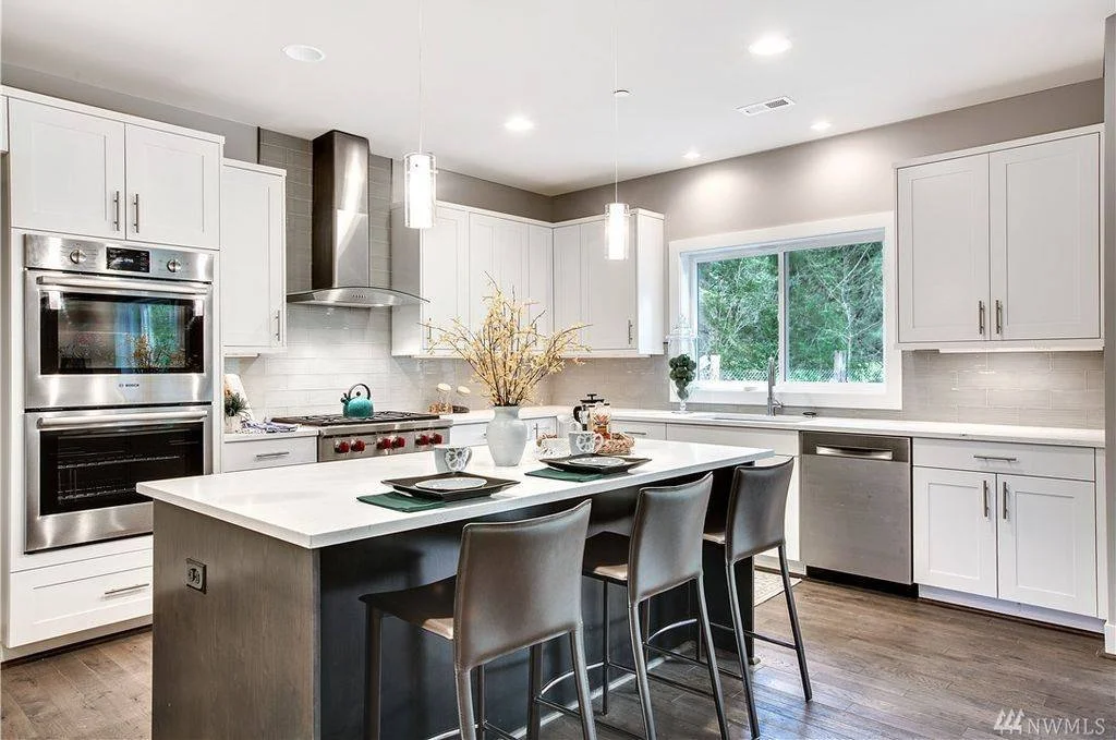 Modern kitchen with white cabinets and stainless steel appliances, including oven, microwave, and stove, a large central island with four barstools, and a window showing greenery outside.