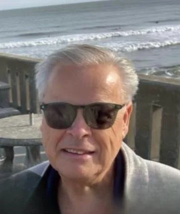 A smiling older man wearing sunglasses and a gray jacket, standing on a beach with the ocean and waves in the background.