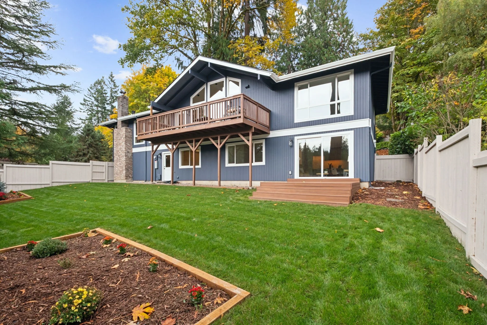 A two-story blue house with white trim and a wooden balcony, set in a landscaped yard with a green lawn, mulch flower bed, and trees in the background.