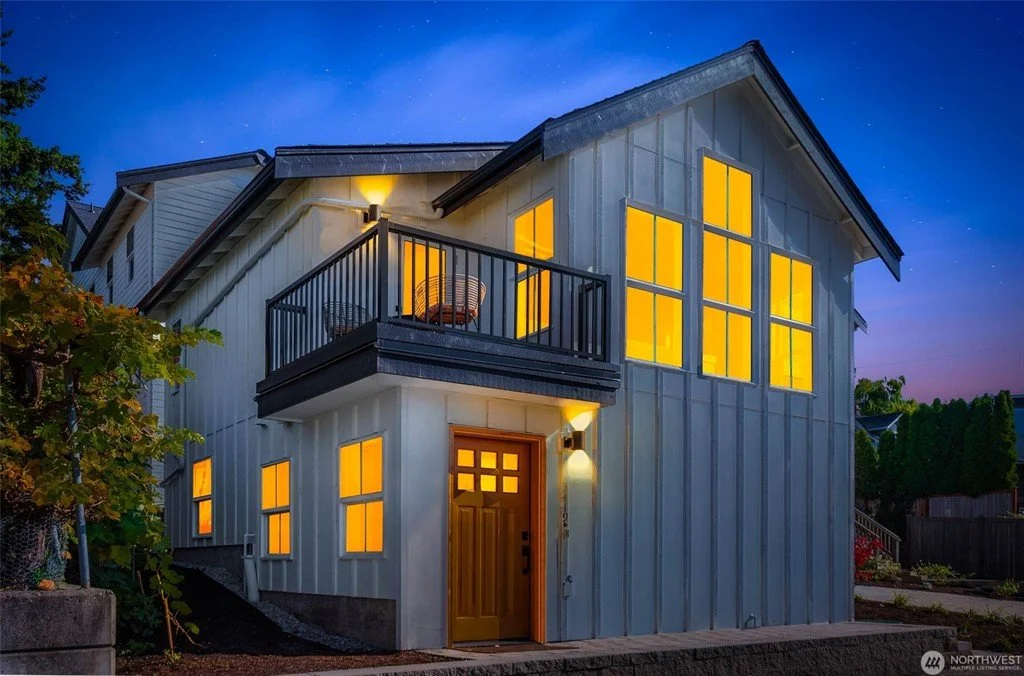 A two-story house with glowing yellow-lit windows and a small balcony, taken during twilight or early night.