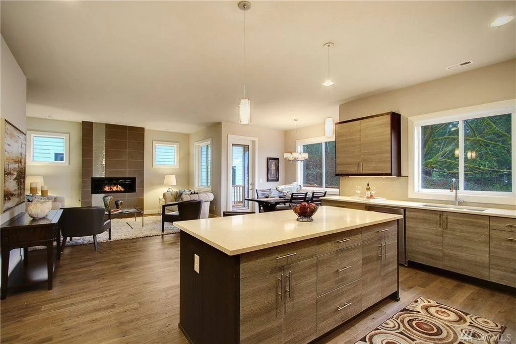 Open-concept living room and kitchen with hardwood floors, modern cabinets, a fireplace, and large windows showing greenery outside.
