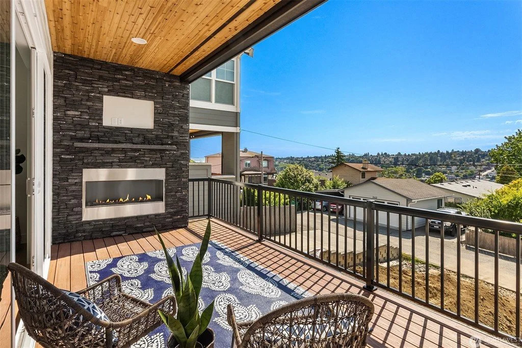 Balcony with black metal railing overlooking residential neighborhood under clear blue sky, with an outdoor fireplace and wicker chairs.