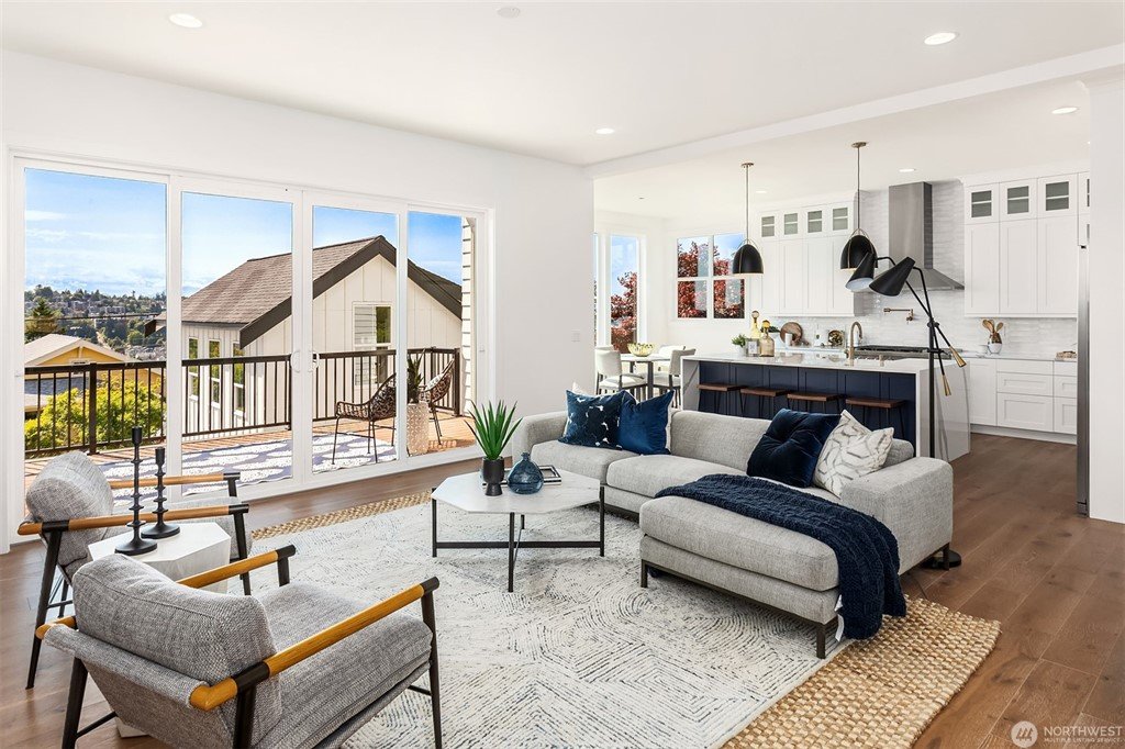 Bright, open living room with large sliding glass doors leading to a balcony, modern gray sofa with blue and white pillows, two armchairs, a rug, a round coffee table, and a kitchen in the background with white cabinets and a navy island.