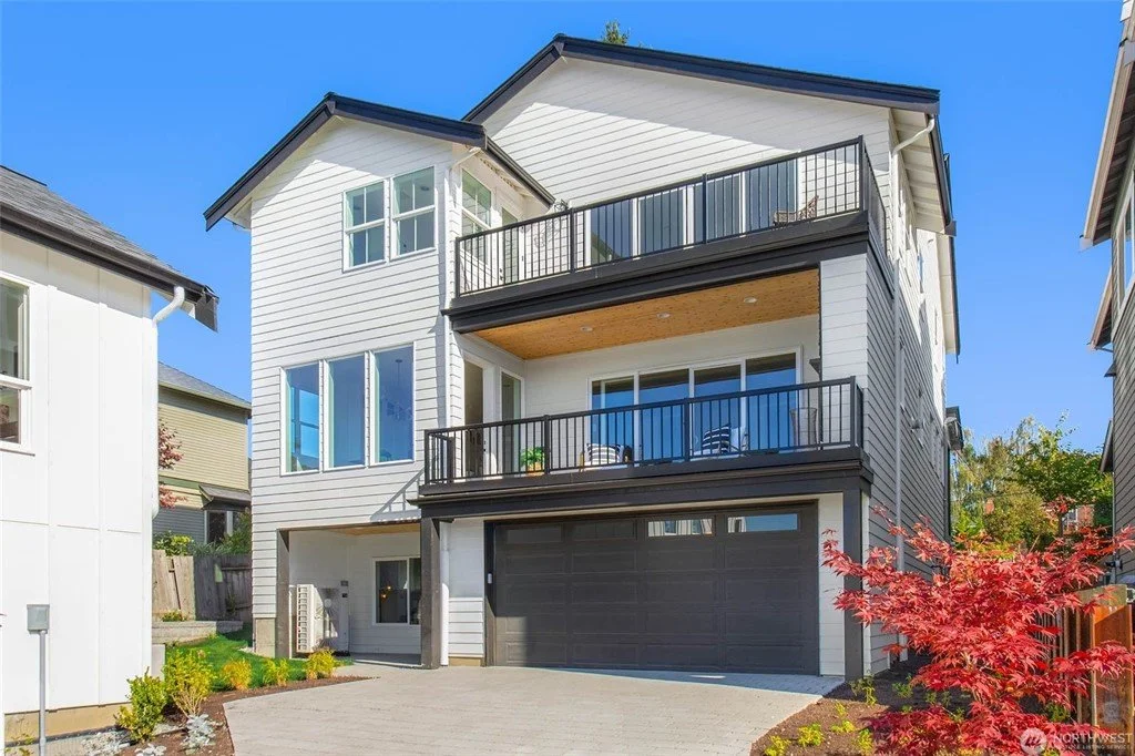 Modern multi-story house with white siding, black accents, two balconies with railing, large windows, and a gray garage door, set against a bright blue sky with some trees and colorful shrubs in the front yard.