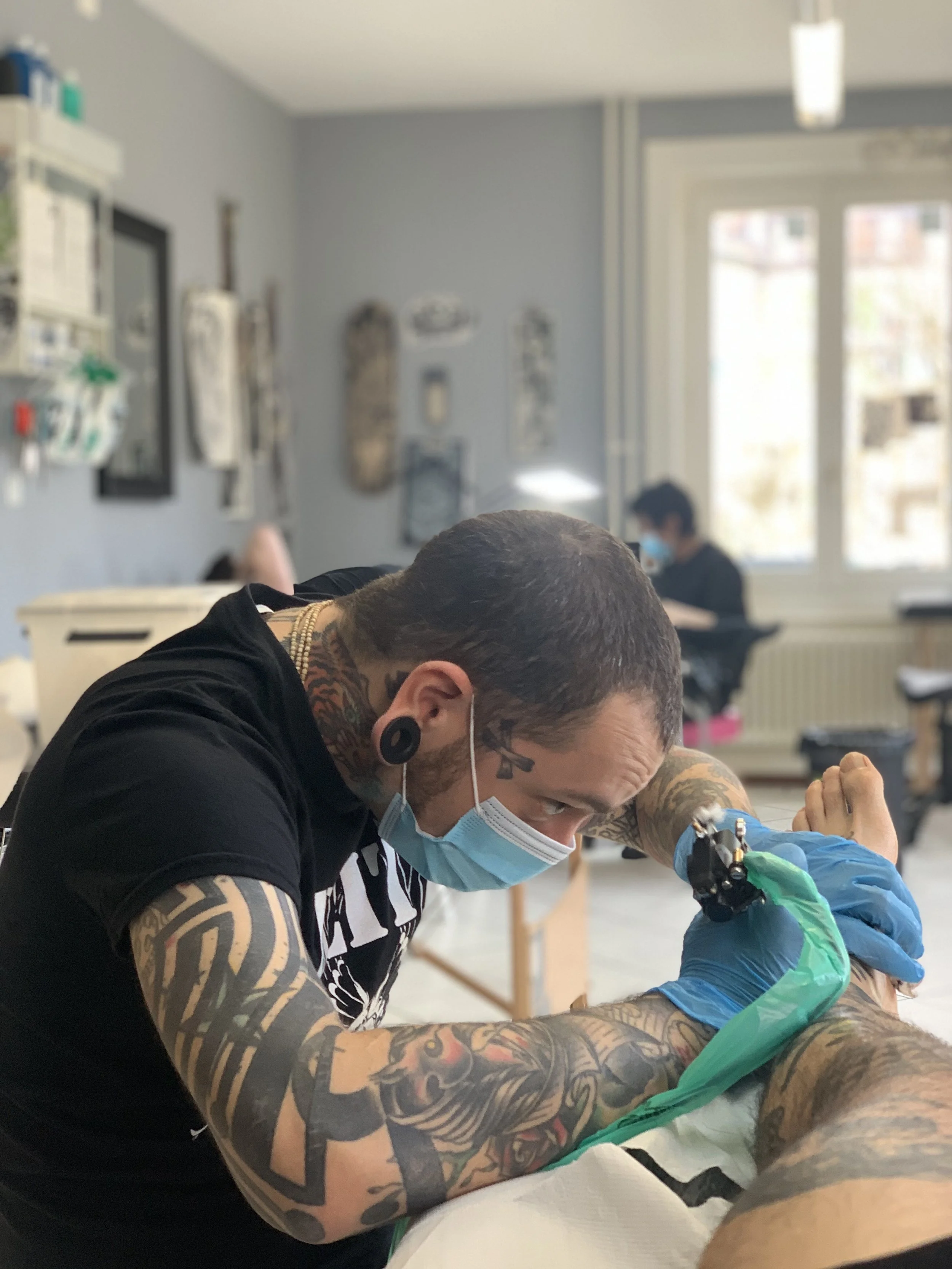 A tattooed tattoo artist wearing a face mask, gloves, and ear gauges is tattooing a person lying on a table inside a tattoo studio. The studio has light blue walls, framed artwork, and large windows in the background.