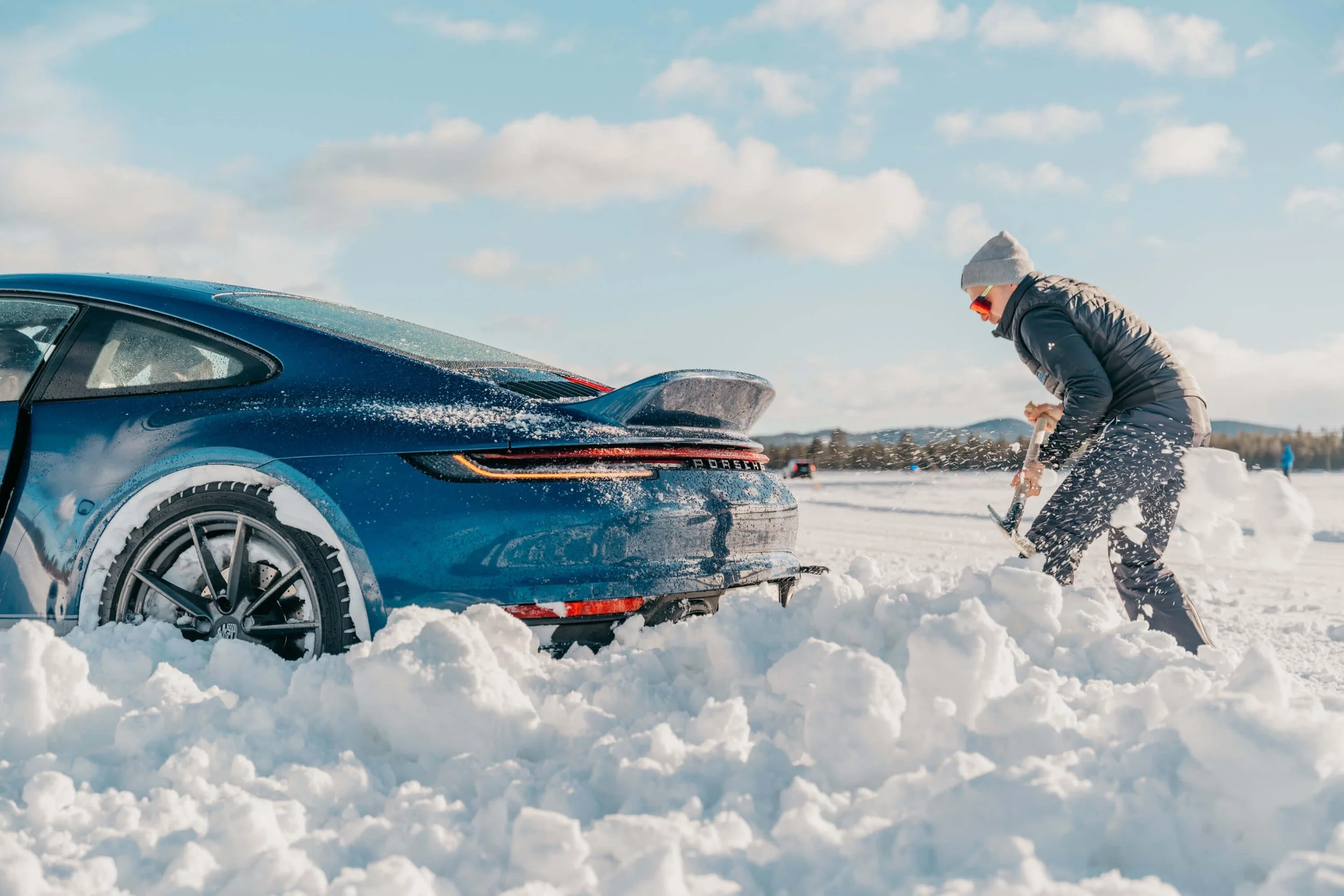 Person digging a Porsche out of deep snow on a frozen lake.